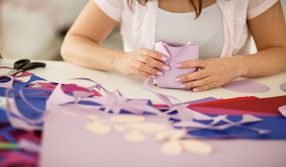 Midsection of unrecognizable female sitting at her workstation and constructing a paper flower out of individual petals she cut out and crafted.