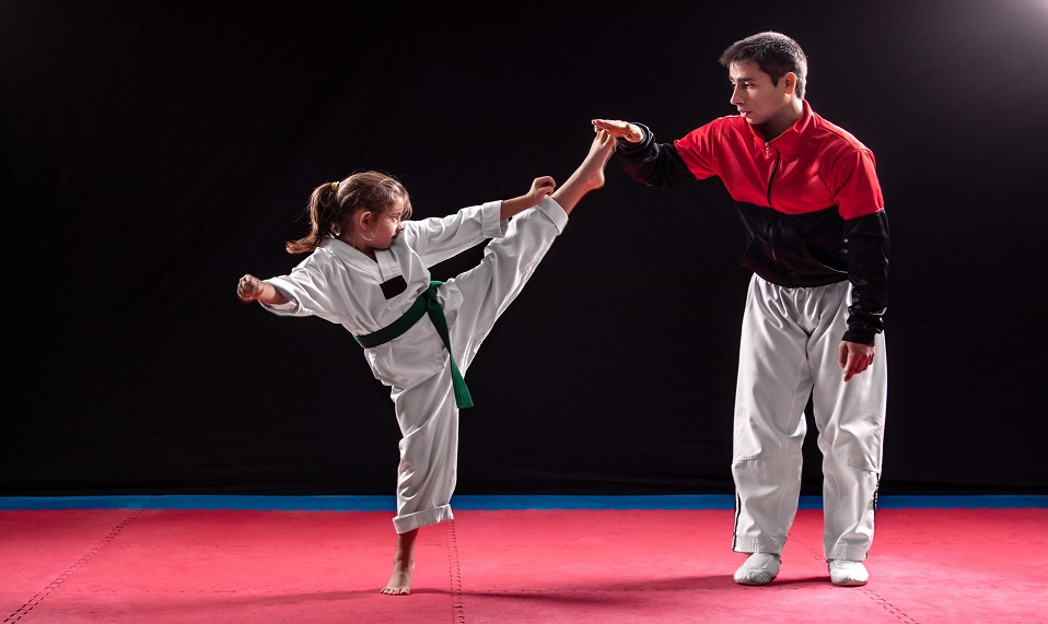 Sports trainer teaching little girl taekwondo and girl trying to make high kick. Taekwondo is one of the most popular martial arts. Theay are working in gym with dark background